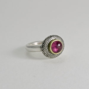 fuscia pink oval tourmaline cabochon set in gold on a stippled oxidised silver oval pebble on slim silver band